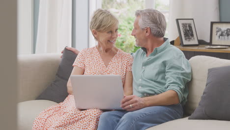 senior retired couple sitting on sofa at home shopping or booking holiday on laptop