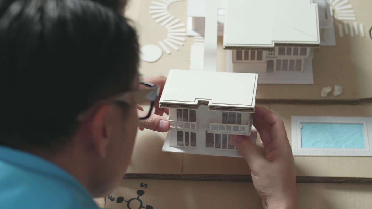 Close Up Of A Man Engineer Looking At Paper Model Of House At The Office, Over Shoulder
