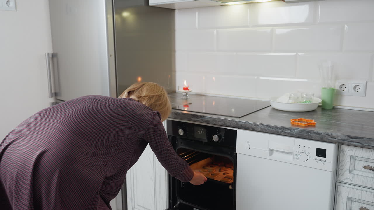mujer colocando galletas en la bandeja de horneado en el horno, con equipos de cocina como rodillo, cortador de galletas y una vela en la mesa en el fondo, preparándose para la sesión de hornear en la cocina