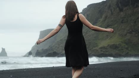 joven hermosa en vestido negro bailando girando en la playa de arena negra islandia, cámara lenta, ondas dramáticas paisaje marino