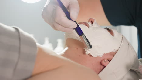 aesthetician spreads thick face scrub in gentle circular motions with soft silicone brush from chin to forehead down to neck of spa visitor under bright lamp for rejuvenating treatment session