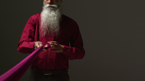 A Sikh man with a long white beard and a turban is wearing a red shirt and holding a purple fabric.