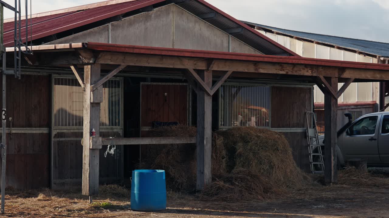Wide exterior shot of a functional wooden stable with horse stalls. Large piles of hay sit under a sheltered porch next to farm equipment and a parked truck