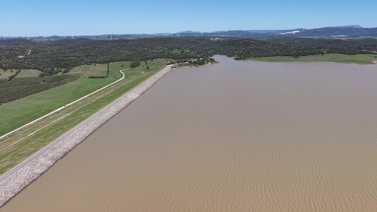 Un plan de drone en suivi du vaste réservoir Embalse del Barbate avec des éoliennes visibles à l'horizon lointain sous un ciel clair.