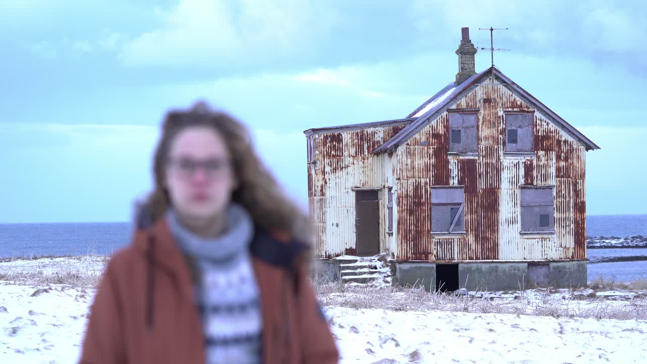 Camera switches focus between rusty house and young girl in a wintery, cold landscape. Closeup stable shot, on 90mm.