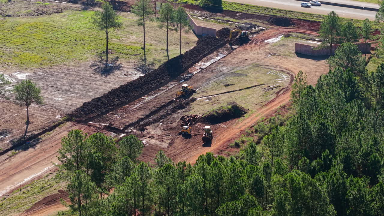 Aerial view captures heavy machinery reshaping sunlit terrain between pine forests and highway. Ideal for infrastructure or environmental documentaries.