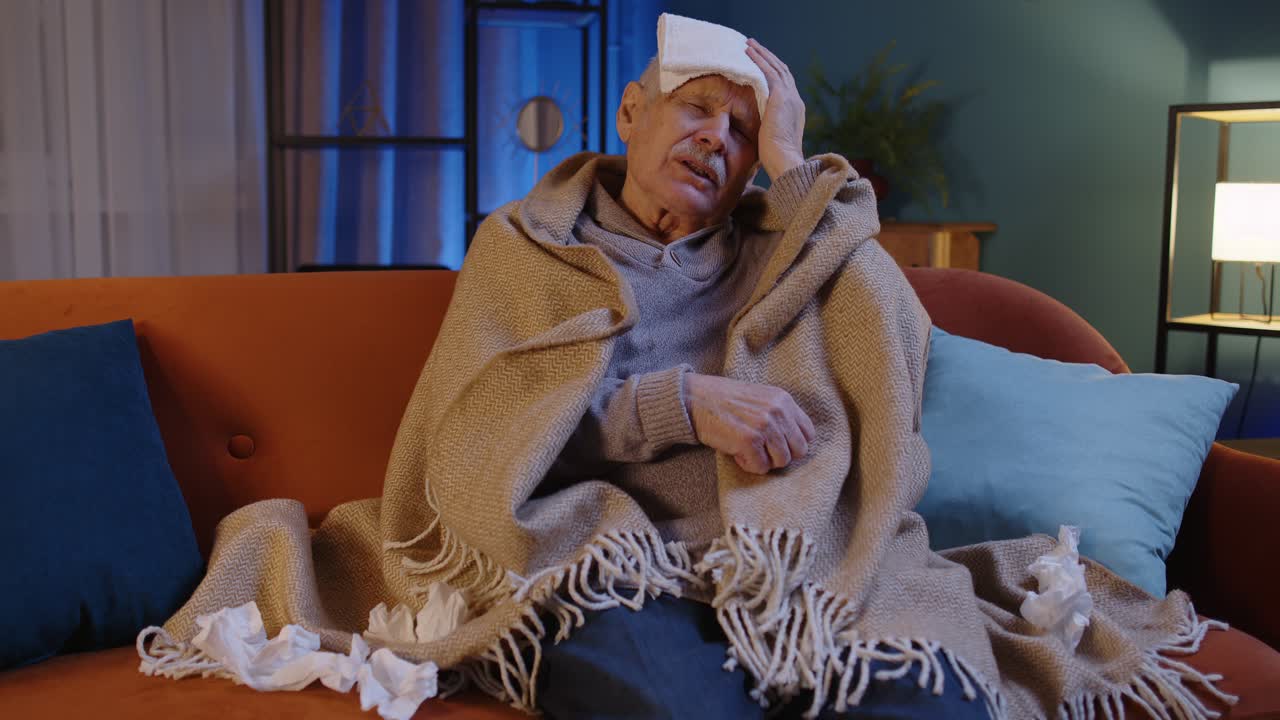 Senior grandfather man suffering from cold or allergy blows nose snot into napkin sitting at home