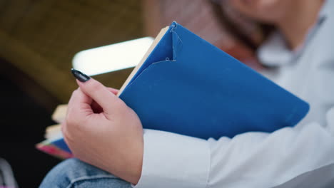 close-up of adult with black polished nails holding blue book on leg, carefully flipping to another page, face slightly blurred with soft bokeh light in background