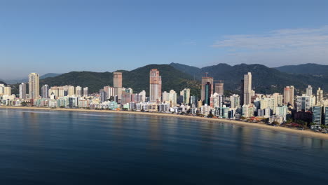 backward-moving drone clip reveals itapema’s towering buildings along meia praia’s coastline, captured under clear blue afternoon skies. ideal for tourism and urban development content