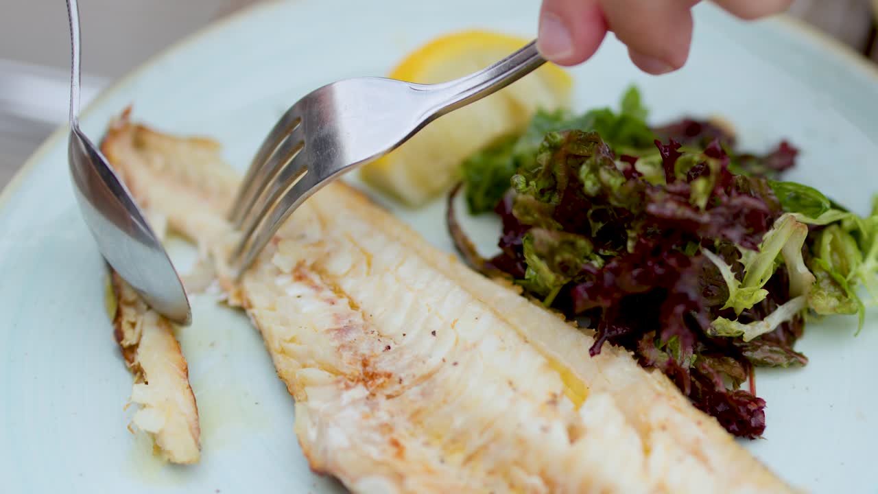 Hand uses fork and spoon to cut grilled fish fillet beside salad under natural daylight