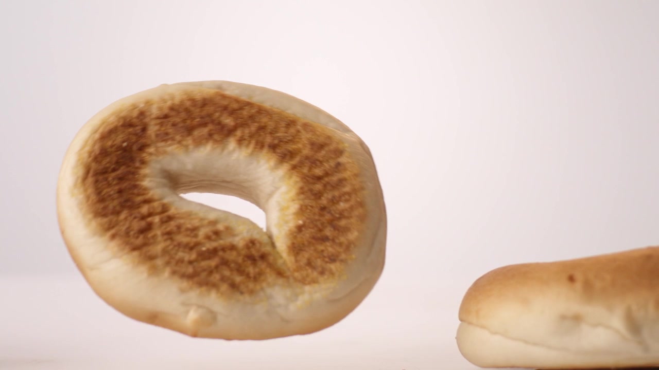 Bagel dorati e gommosi che rimbalzano e rotolano su uno sfondo bianco in uno scatto macro da studio al rallentatore. Contenuti per gastronomie newyorkesi, branding per negozi di bagel, reel per menu brunch.