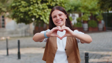 portrait of caucasian young woman makes symbol of love showing heart sign to camera on city street