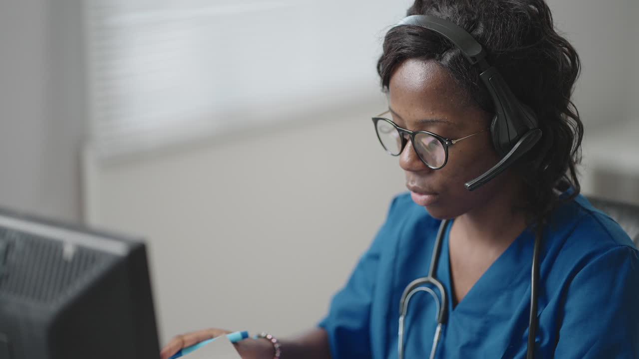 A black woman doctor wearing headphones sits at a table with a computer and takes calls from patients looks at their medical records and enters them into the clinic schedule