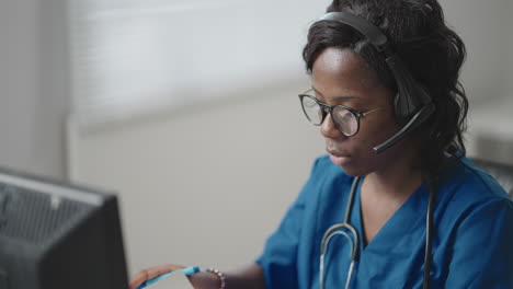 una mujer negra que usa auriculares se sienta en una mesa con una computadora y recibe llamadas de pacientes mira sus registros médicos y los ingresa en el horario de la clínica