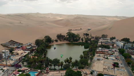 toma aérea que se eleva sobre el pequeño pueblo de huacachina en perú, mostrando el oasis de agua rodeado por el desierto