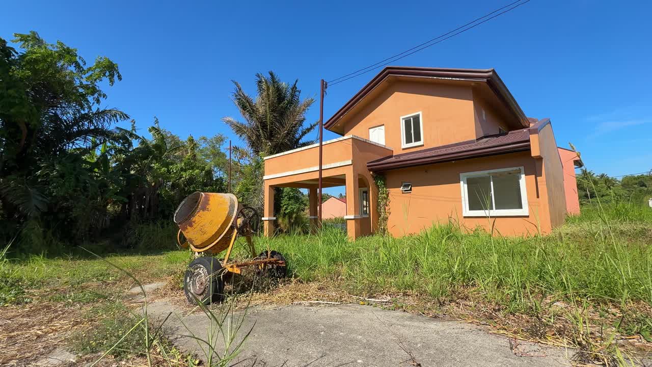 Sudut pandang samping dari permukaan tanah sebuah rumah yang sudah selesai, tidak berpenghuni, dengan rumput tumbuh hingga ke dinding dan peralatan konstruksi tua di sampingnya