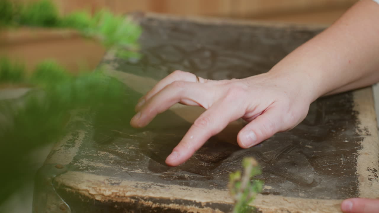 close hand view of person drawing simple shape with finger on dusty old box surface while soft green leaf and round yellow bokeh blur appear in foreground creating calm creative moment indoors