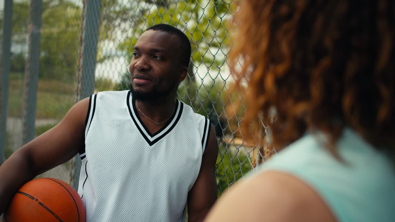 Two men are talking about basketball: a man with Black skin in a white T-shirt holds a basketball in his hand and communicates with his friend, a curly red-haired man