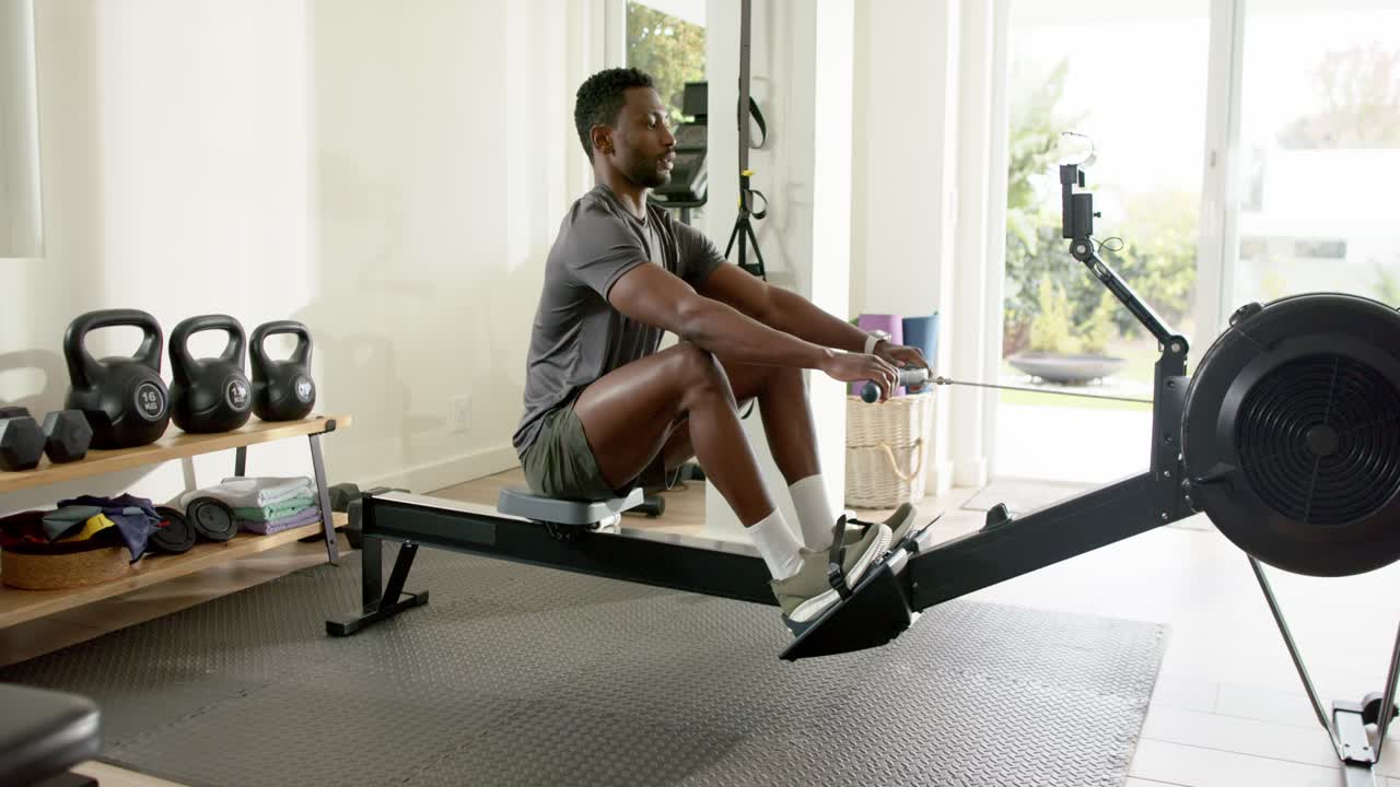 Mid adult African American man glancing at smartphone to track workout, rowing on machine at home