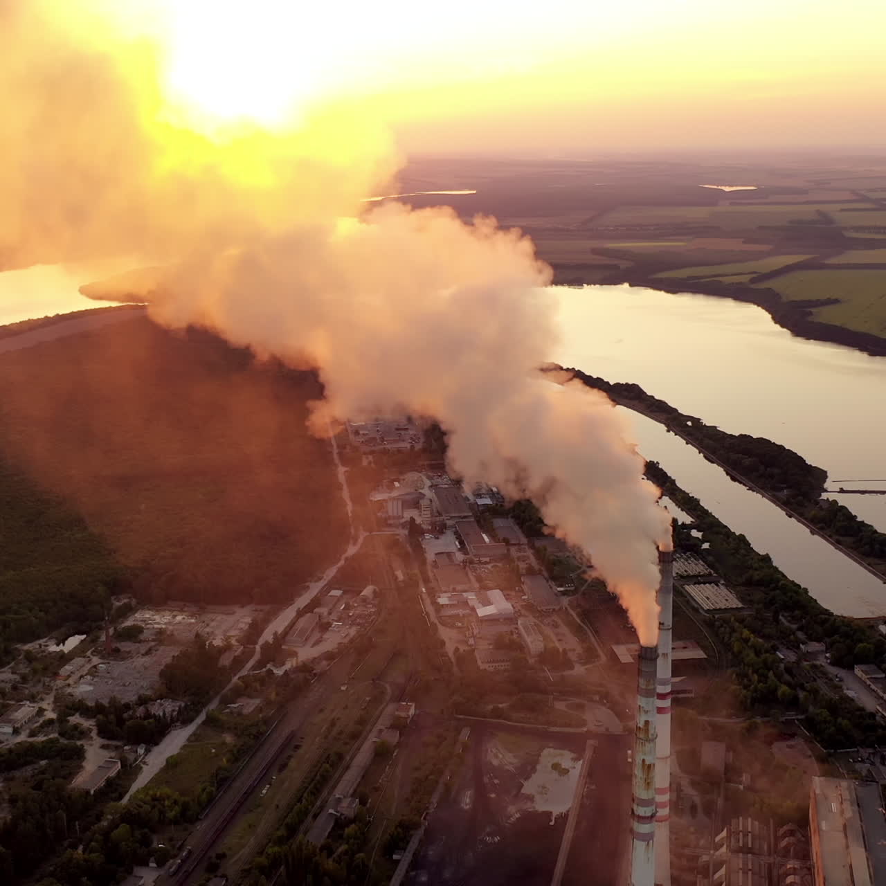 Aerial view of a factory among nature. Industrial pipes pollute the atmosphere with smoke at sunset. Beautiful river next to the huge metallurgy plant.