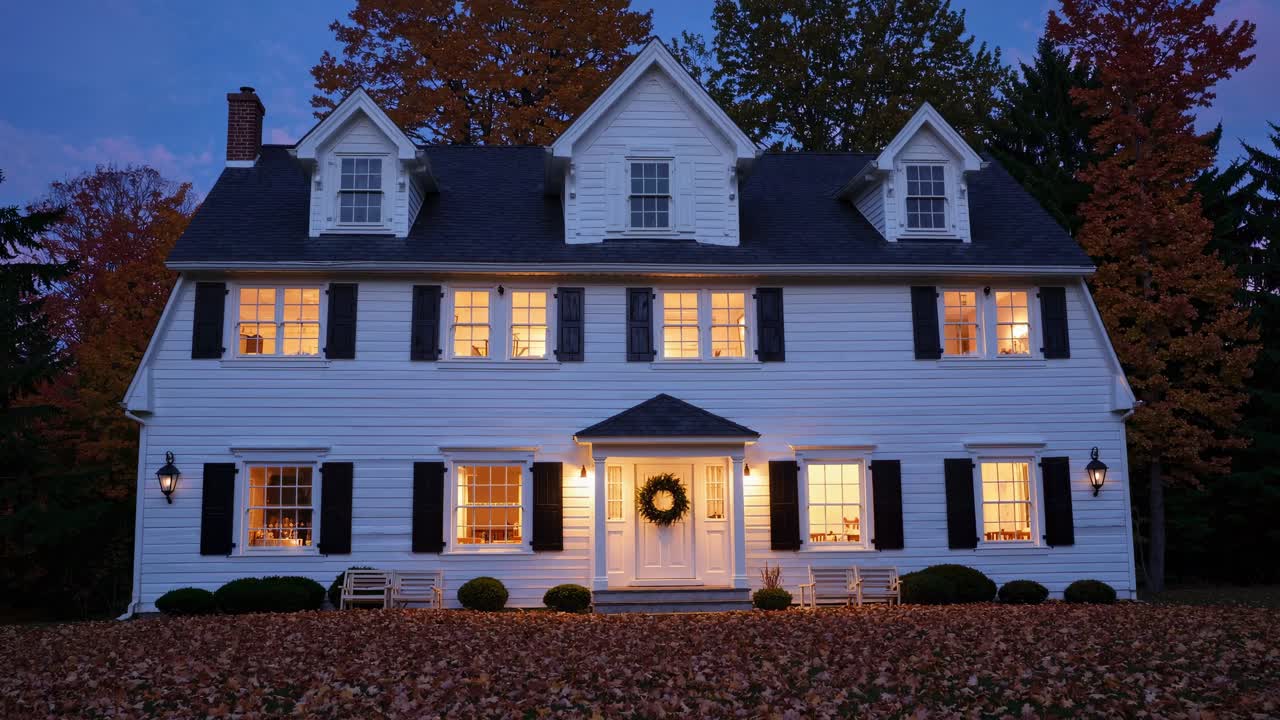 Warm lights glowing from the windows of a large white classic American house decorated with a Christmas wreath at the front door, surrounded by colorful autumn leaves at twilight