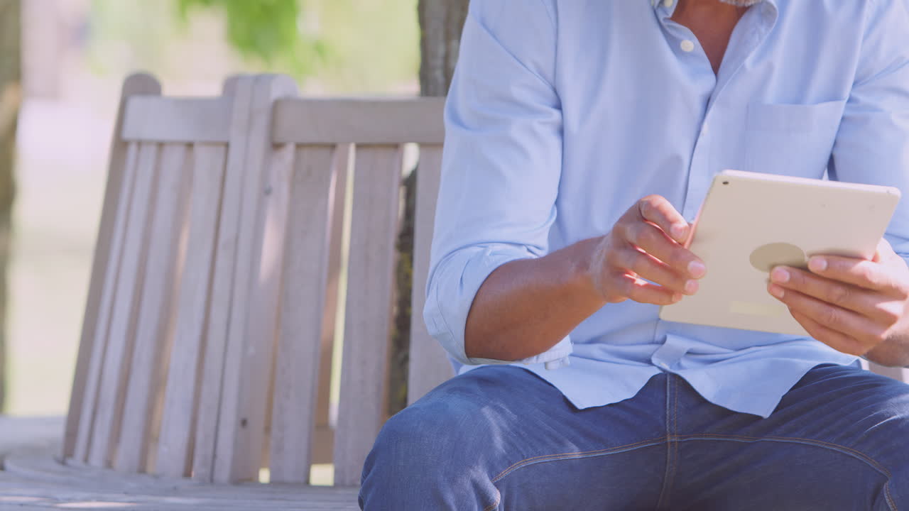 primer plano de un hombre sentado en un banco bajo un árbol en un parque de verano usando una tableta digital