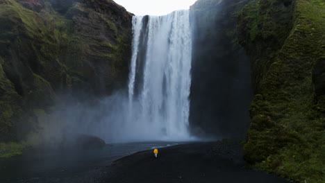 Man-In-Yellow-Jacket-Walking-Near-Skogafoss-Waterfall-In-Iceland---Drone-Shot