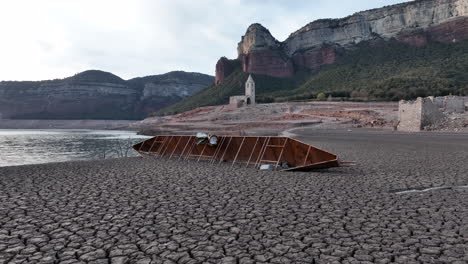 Shipwreck-of-wooden-boat-on-dry-cracked-muddy-shores-of-Sau-marsh-with-church-ruin-in-background,-Catalonia,-Spain