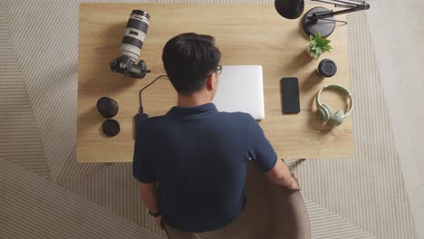 top view of a male editor walking into the workspace sitting down and using a laptop next to the camera editing photo of a female at home