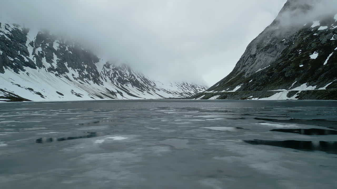 Drone shot of a frozen lake in Norway. There are still patches of ice on the lake. The mountains and low-hanging clouds are reflected in the water's surface