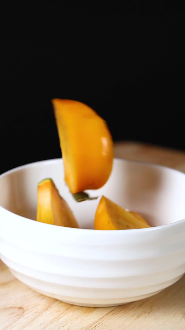 Fresh orange persimmon wedges drop into a white ceramic bowl against a dark background