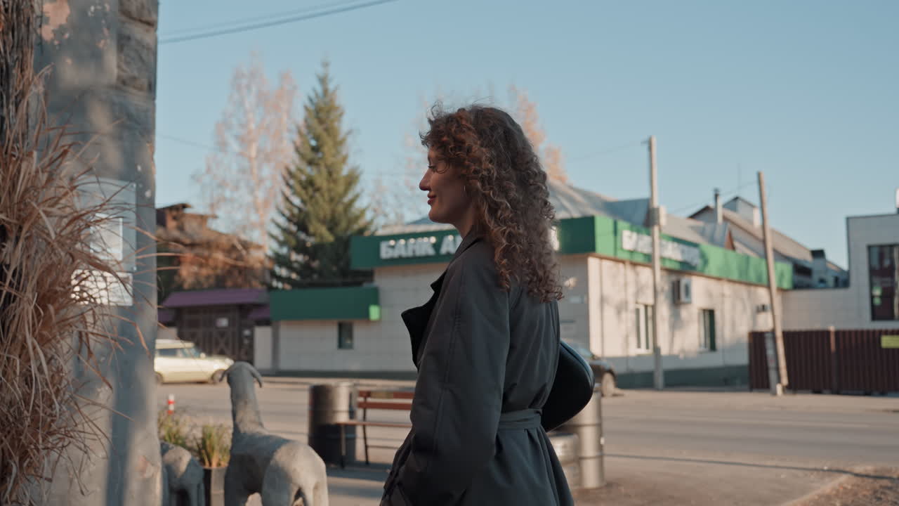 Autumn street florist approaches shop gate, curlyhaired woman in dark trench coat walks along iron fence and opens wooden door into cozy boutique, soft afternoon light, pensive expression, urban