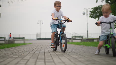 two siblings are seen riding bicycles on a paved walkway, the younger child leads ahead on a smaller bike, while the older sibling follows behind