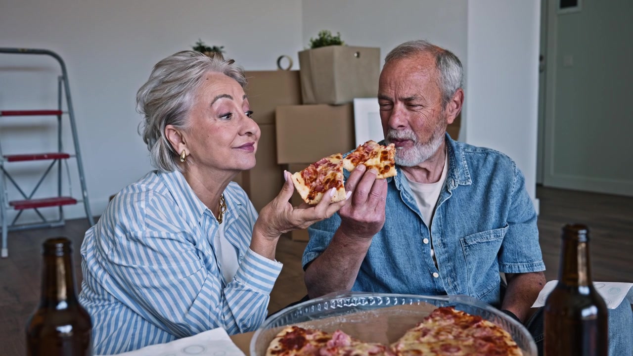 Coppia anziana felice che mangia la pizza nella nuova casa con gli scatoloni del trasloco