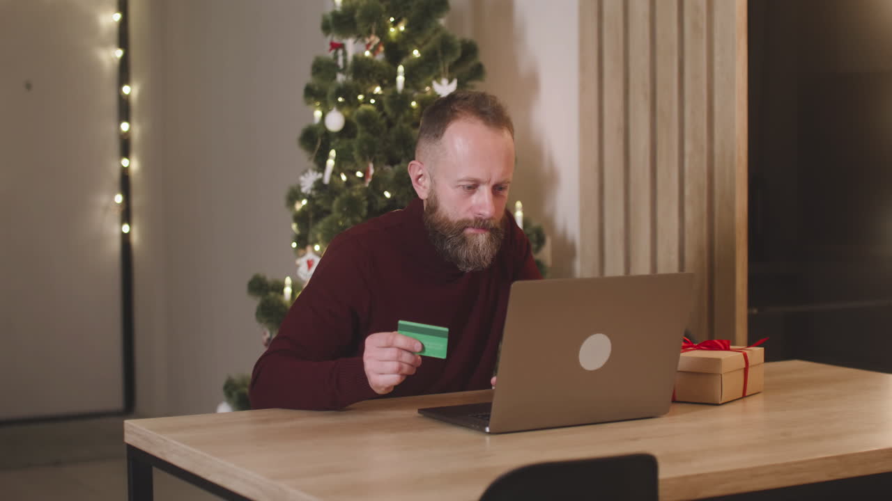 hombre de cabello rojo comprando en línea con una tarjeta de crédito usando una computadora portátil sentada en una mesa cerca de un regalo en una habitación decorada con un árbol de navidad