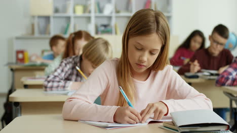 Portrait-Of-The-Teen-Beautiful-Blond-Girl-Writing-In-The-Copybook-At-The-Lesson-In-The-School-And-Smiling-To-The-Camera-Happily