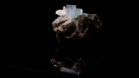 an aquamarine crystal emerging from the matrix of muscovite turns slowly with a reflection of the sample apparent in the glass base