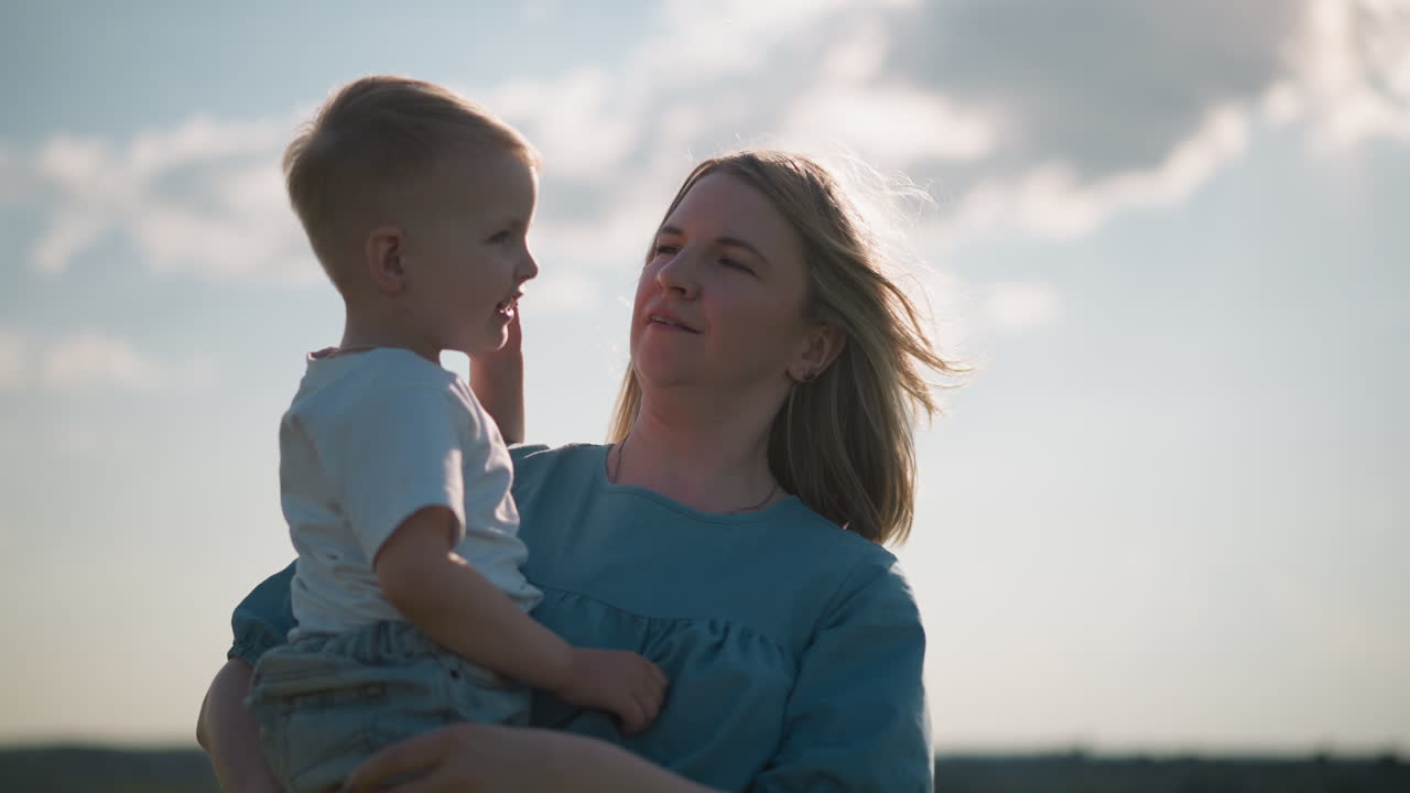 A woman in a blue dress carries her young son, who is wearing a white top, as he smiles brightly. They share a joyful moment outdoors on a sunny day, with soft light illuminating their faces