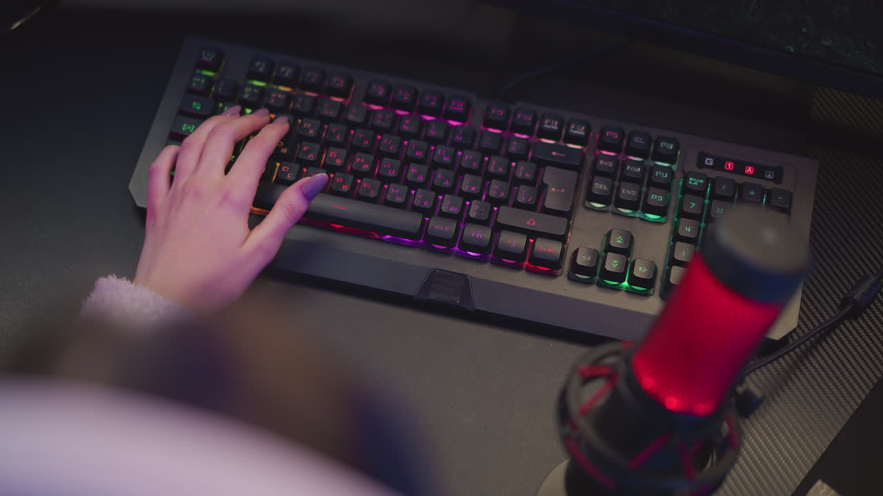 aerial view of female gamer typing on rgb backlit keyboard with polished nails beside red microphone setup on black table surface in illuminated gaming environment