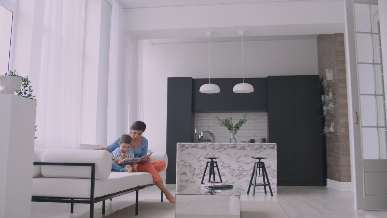 A young mother with a child reading a book sitting in a bright white interior of the house in the living room on the couch