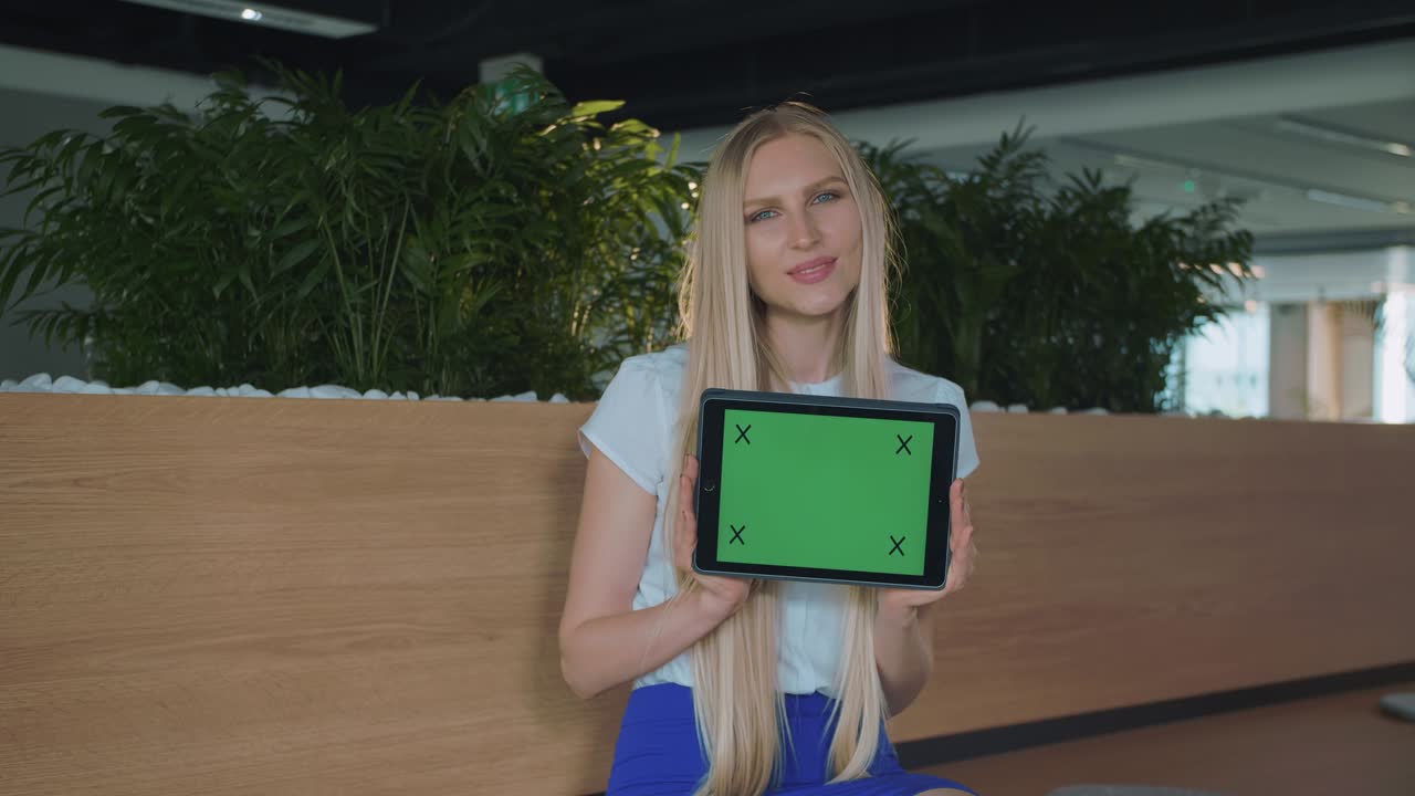 Woman showing tablet in office. Young smiling blond woman showing tablet with green chromakey screen looking at camera and smiling in office