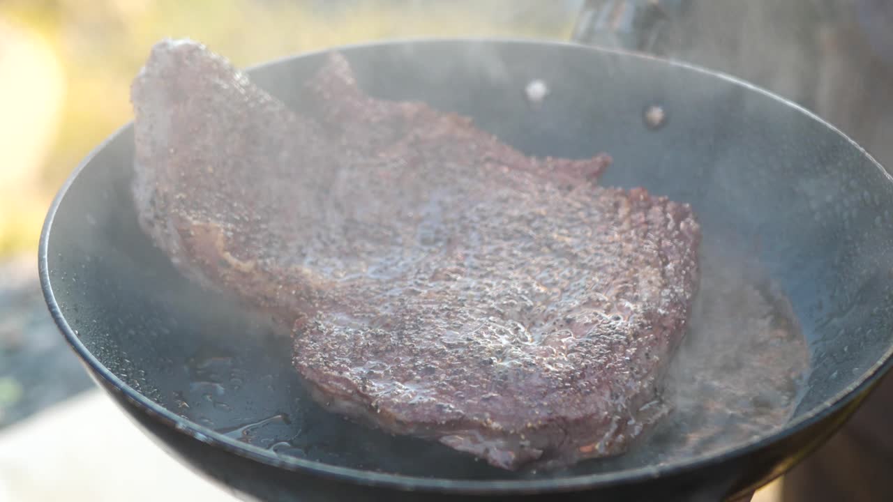 Close up shot of Prime rib entrecôte being flipped and roasted brown in sizzling hot pan, with the aromas condensing and disappearing into a camping outdoor environment