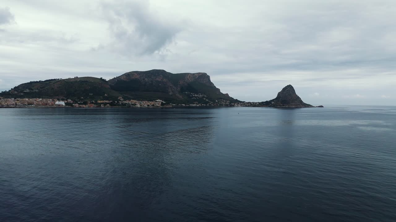 Drone footage of Porticello coastline showing Monte Catalfano and Capo Zafferano across calm blue water under a cloudy sky in Sicily, Italy