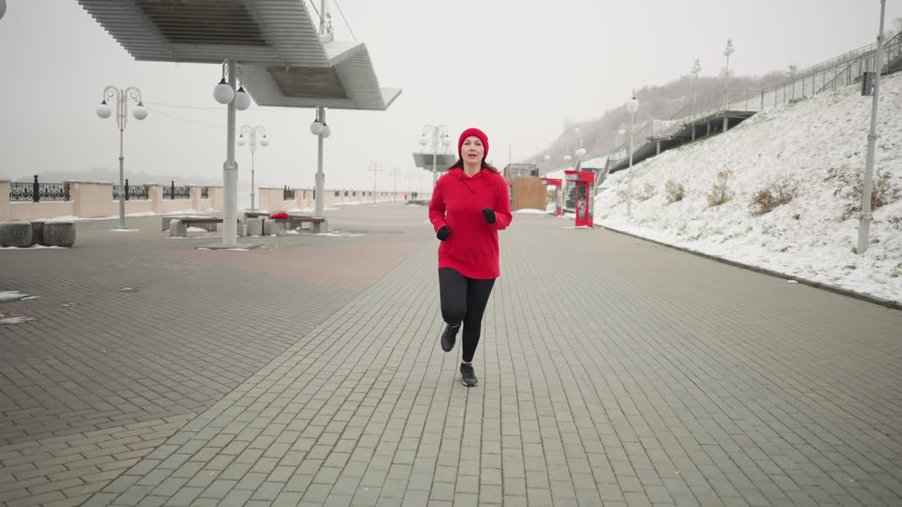 Mom on maternity leave jogging outdoors on interlocked pavement during winter, serene environment with modern urban features like benches, snow, and a snowy hill nearby