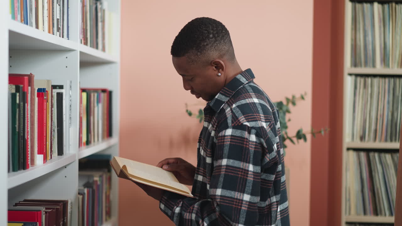 Man reads book standing next to shelf. Thoughtful dark-skinned bookworm dips into literature standing in premise of college library. Reading as hobby