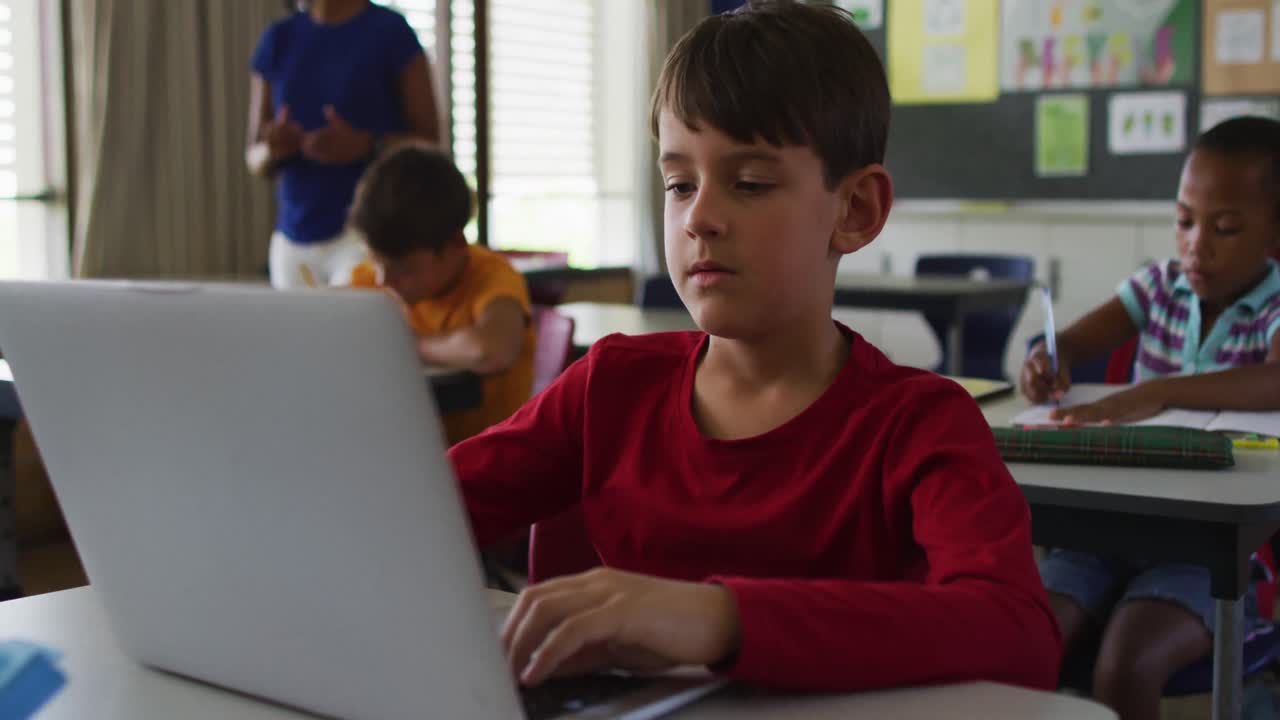 retrato de un escolar caucásico feliz sentado en el aula, usando una computadora portátil, mirando a la cámara