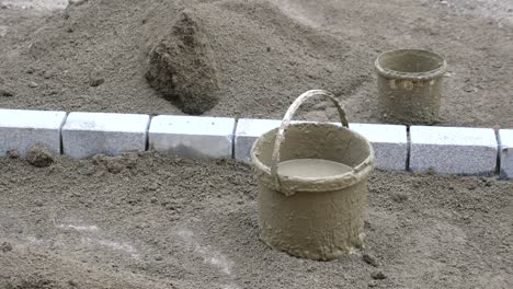 two buckets of concrete sit near a row of grey paving stones on a bed of sand.