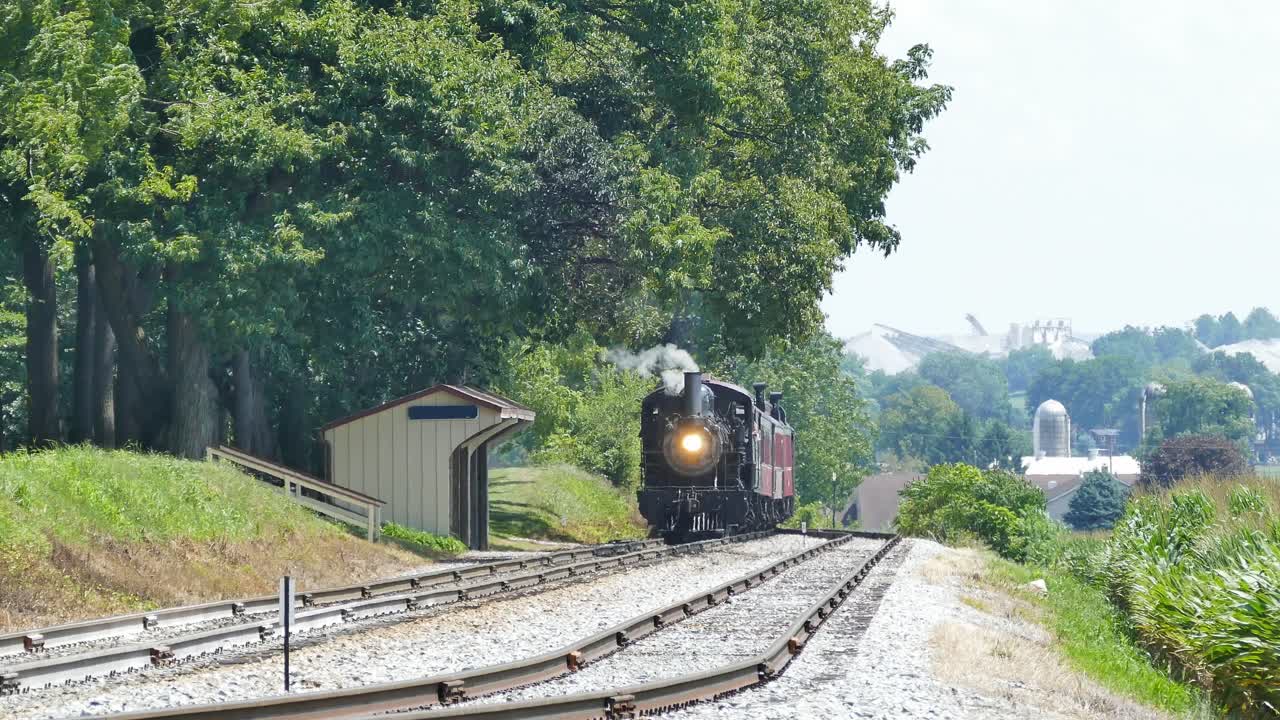 Una locomotora de vapor avanza por las vías en una zona rural verde. Pasa por una pequeña estación rodeada de árboles. El humo se eleva a medida que avanza en un día soleado.