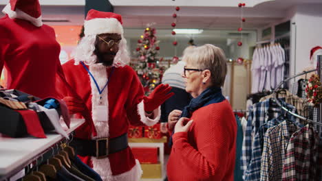 manager wearing santa claus costume around shopping mall fashion boutique chatting with elderly woman. supervisor in holiday themed suit showing senior client red garment piece