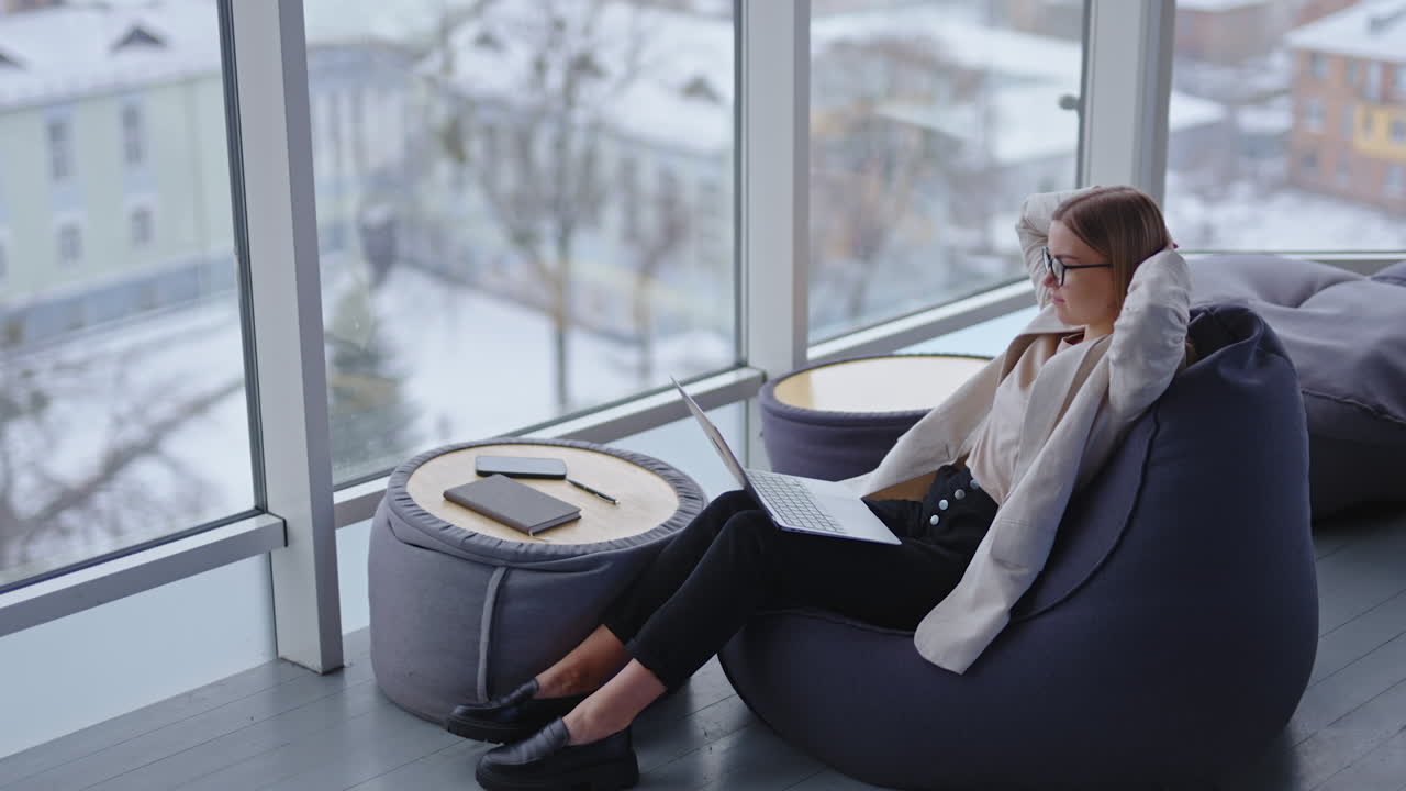 Cozy workplace in modern office. Lady sits at bean bag chair and works on the laptop. Happy lady puts hands behind her hand and sits smiling and relaxing.
